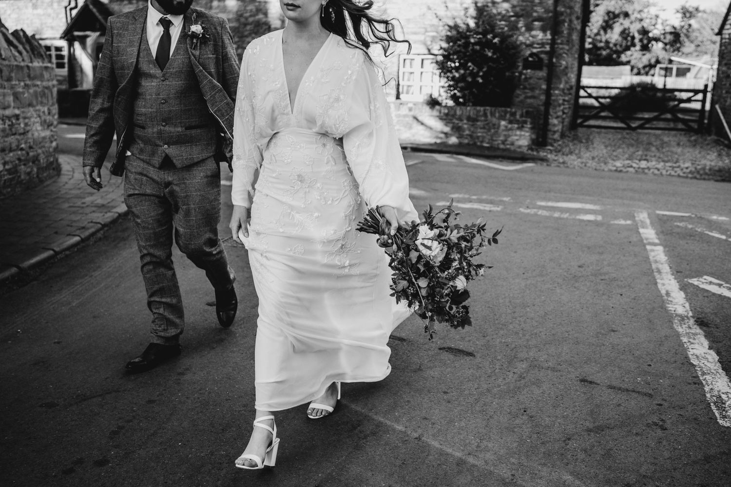 A couple walks down a street after their wedding with the bride holding a floral bouquet.