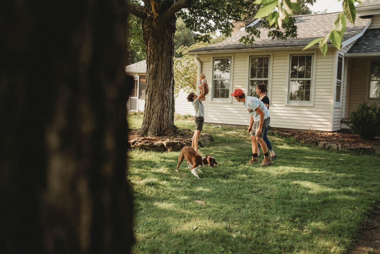 Children and a dog play in a sunny backyard with a white house in the background.