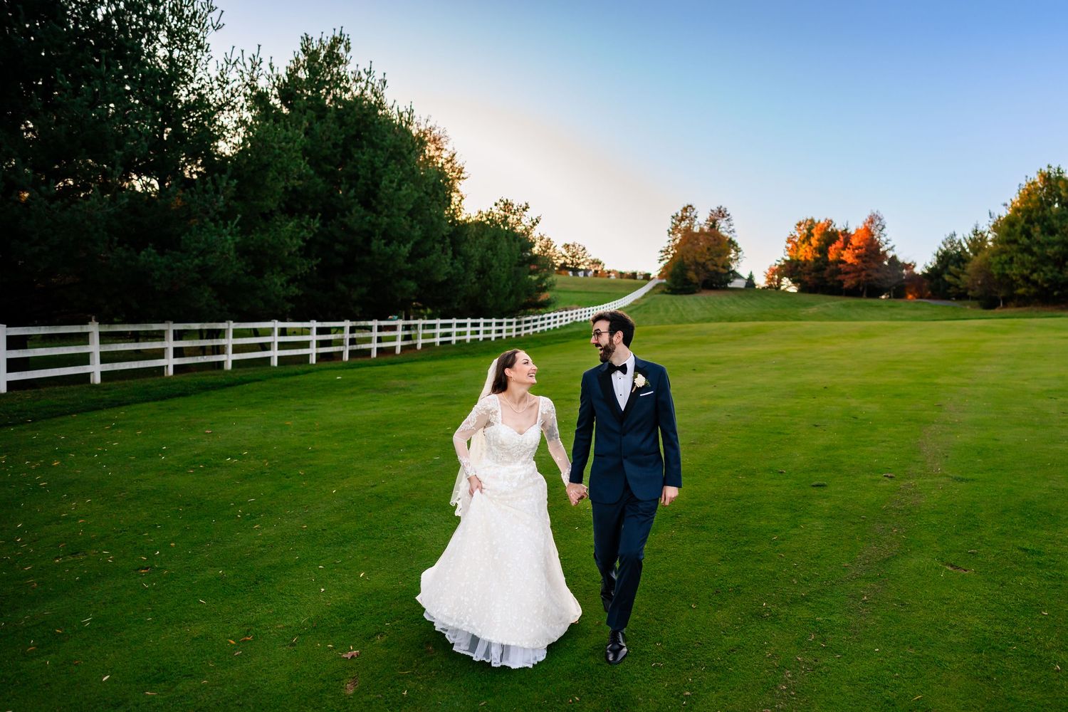 Newlyweds walking along white fence on lush green lawn during golden hour.