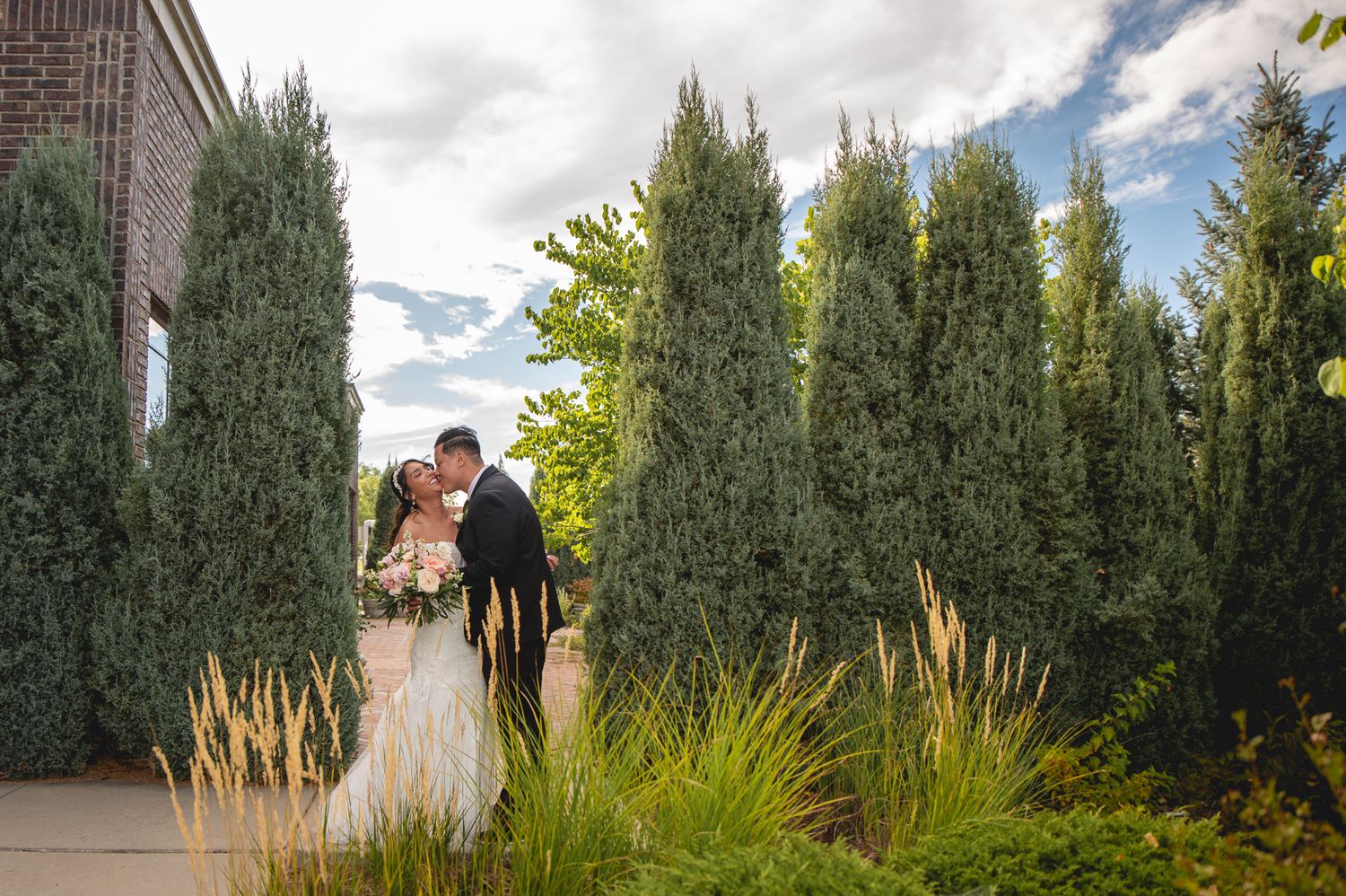 A silhouetted couple stands among tall cypress trees against a cloudy sky.