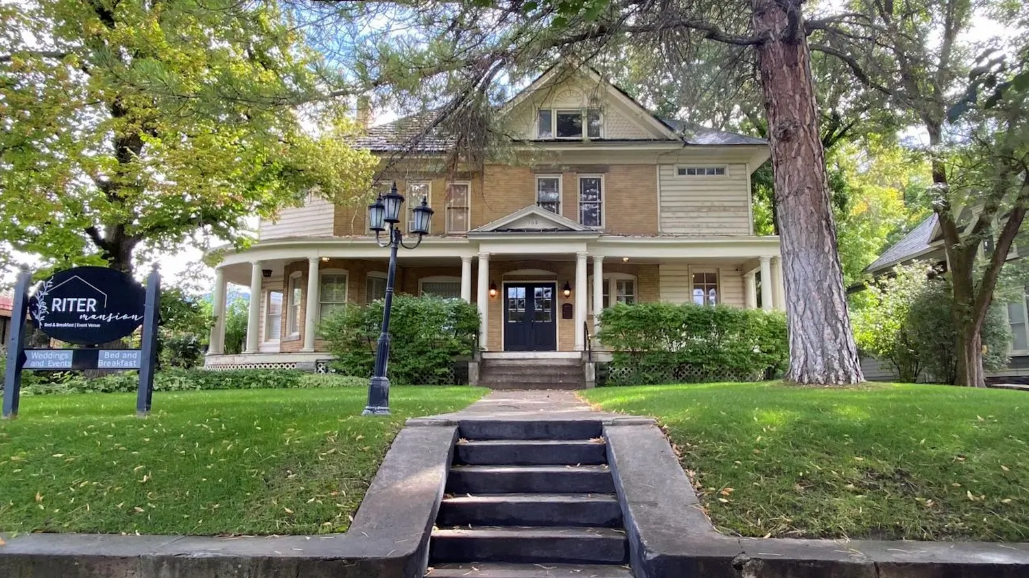Historic yellow mansion with grand staircase entrance and tall trees.