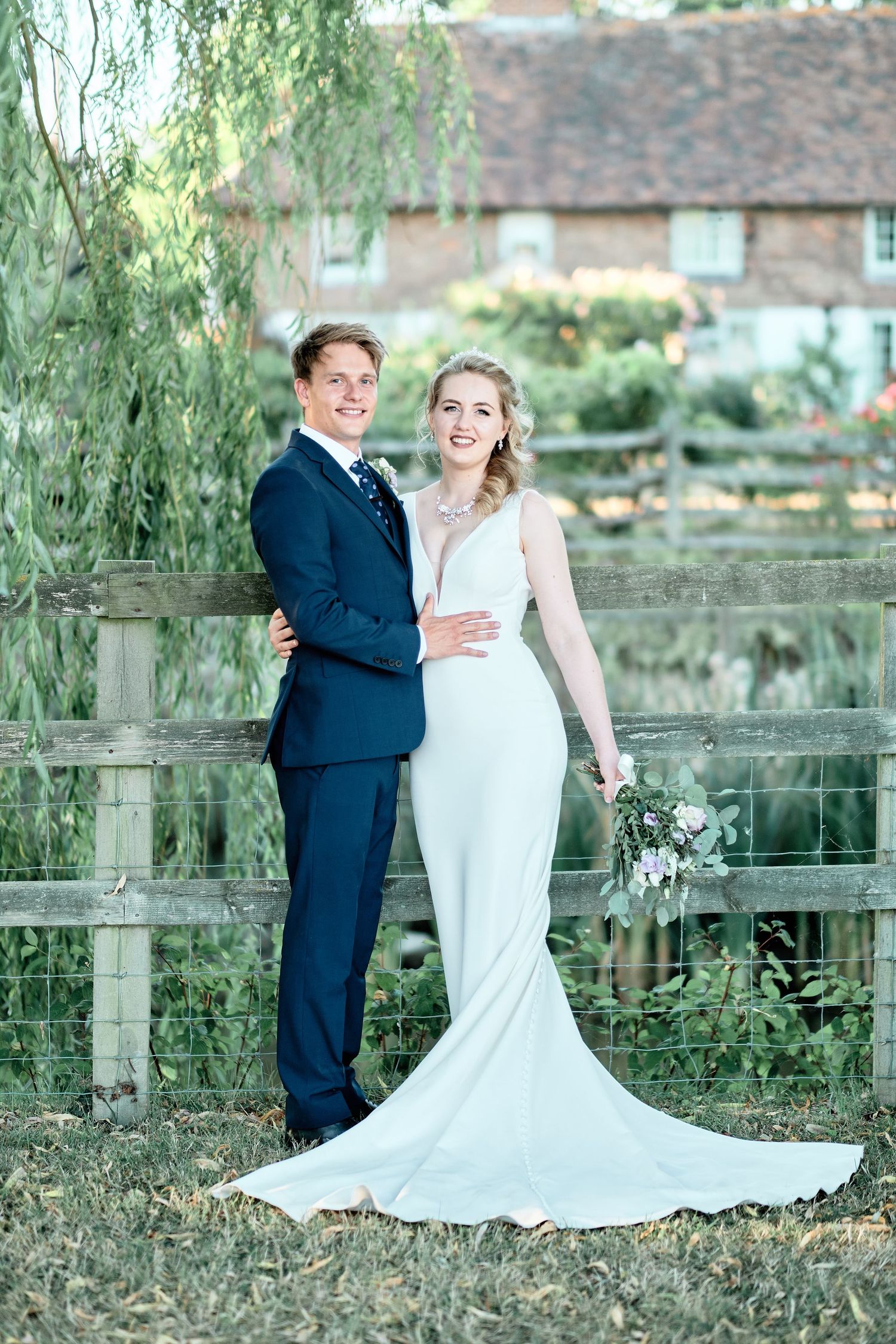 Wedding couple poses by rustic fence with trailing willow trees and country house in background.