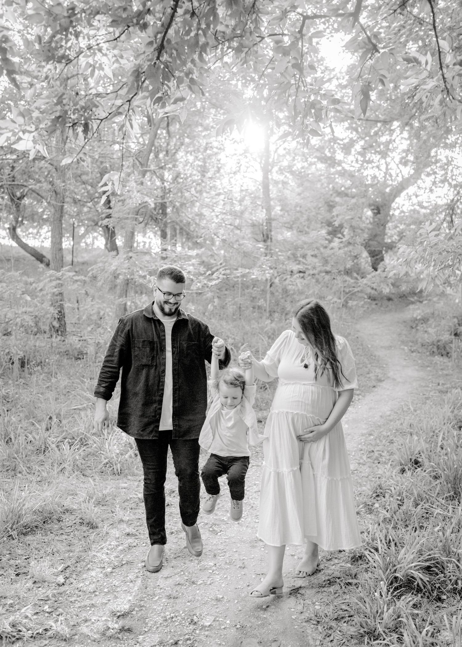 parents swinging their little boy as they all walk hand in hand. mom cradling her baby bump. Boerne, TX