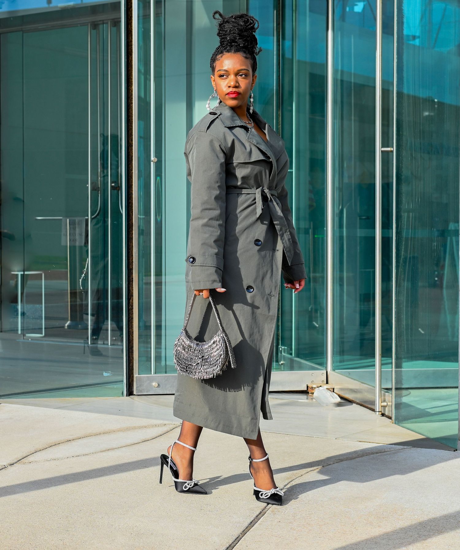 Fashion photo of grey trench coat outfit with heels and handbag against glass building exterior.