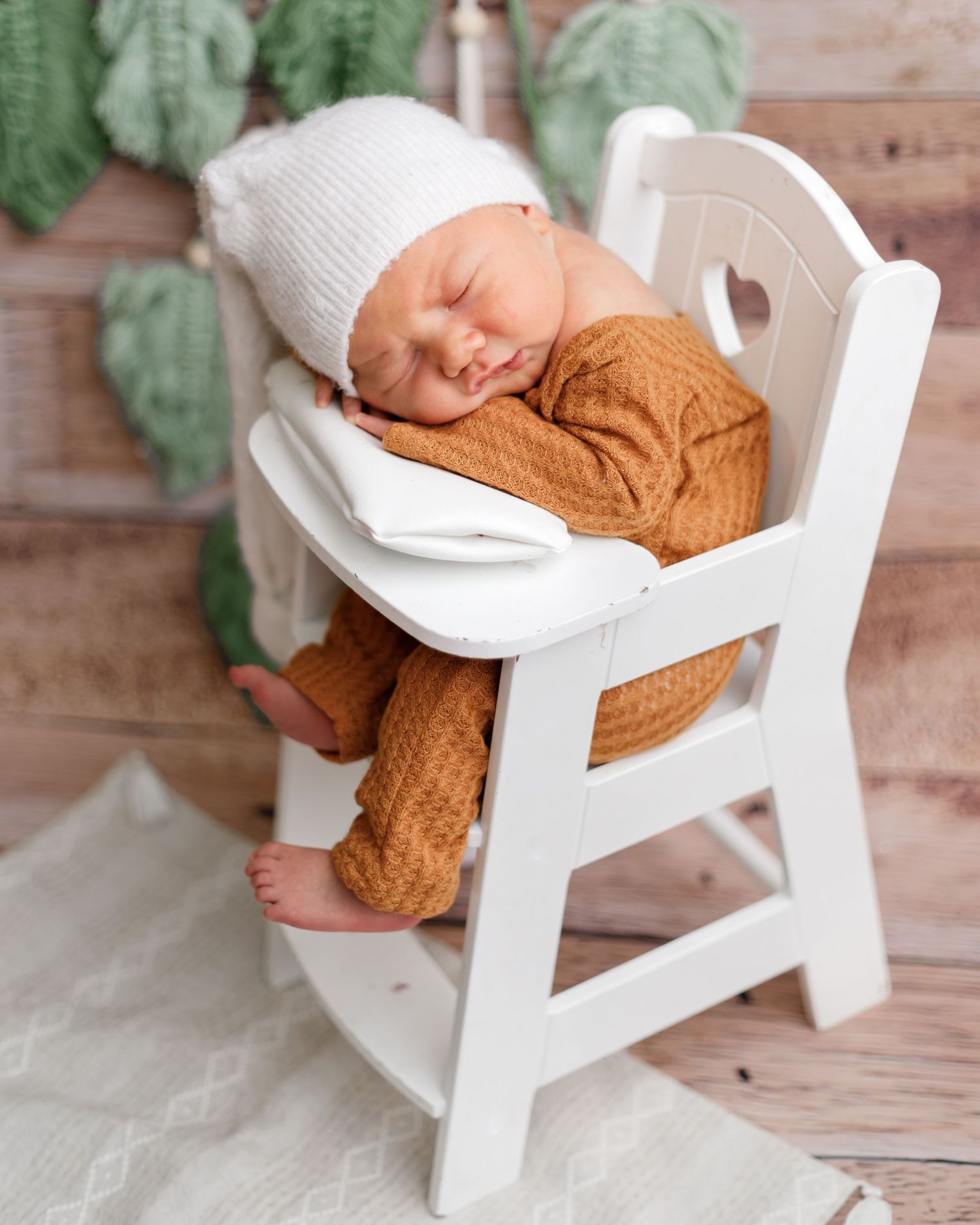 Baby napping in white wooden chair wearing brown outfit and white knit hat.