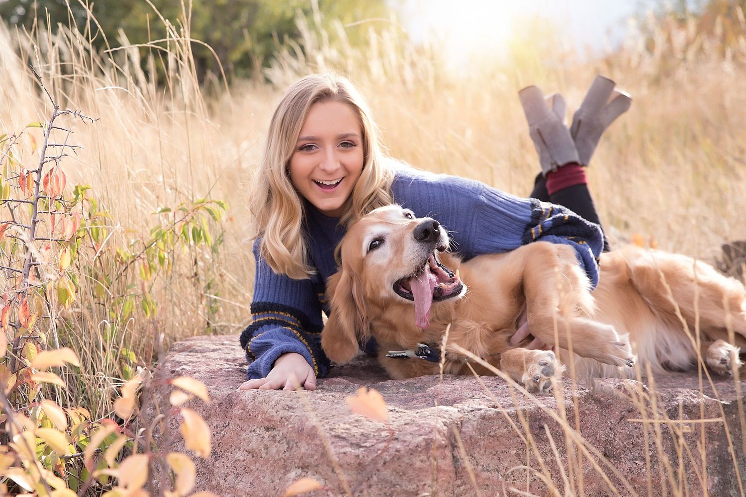A happy person lies on rocks with their golden retriever dog in golden sunlit field.