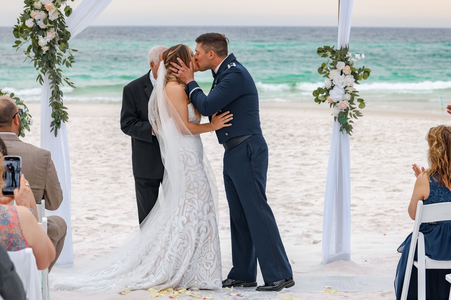 Beach wedding ceremony under floral arch with turquoise ocean waters in background.