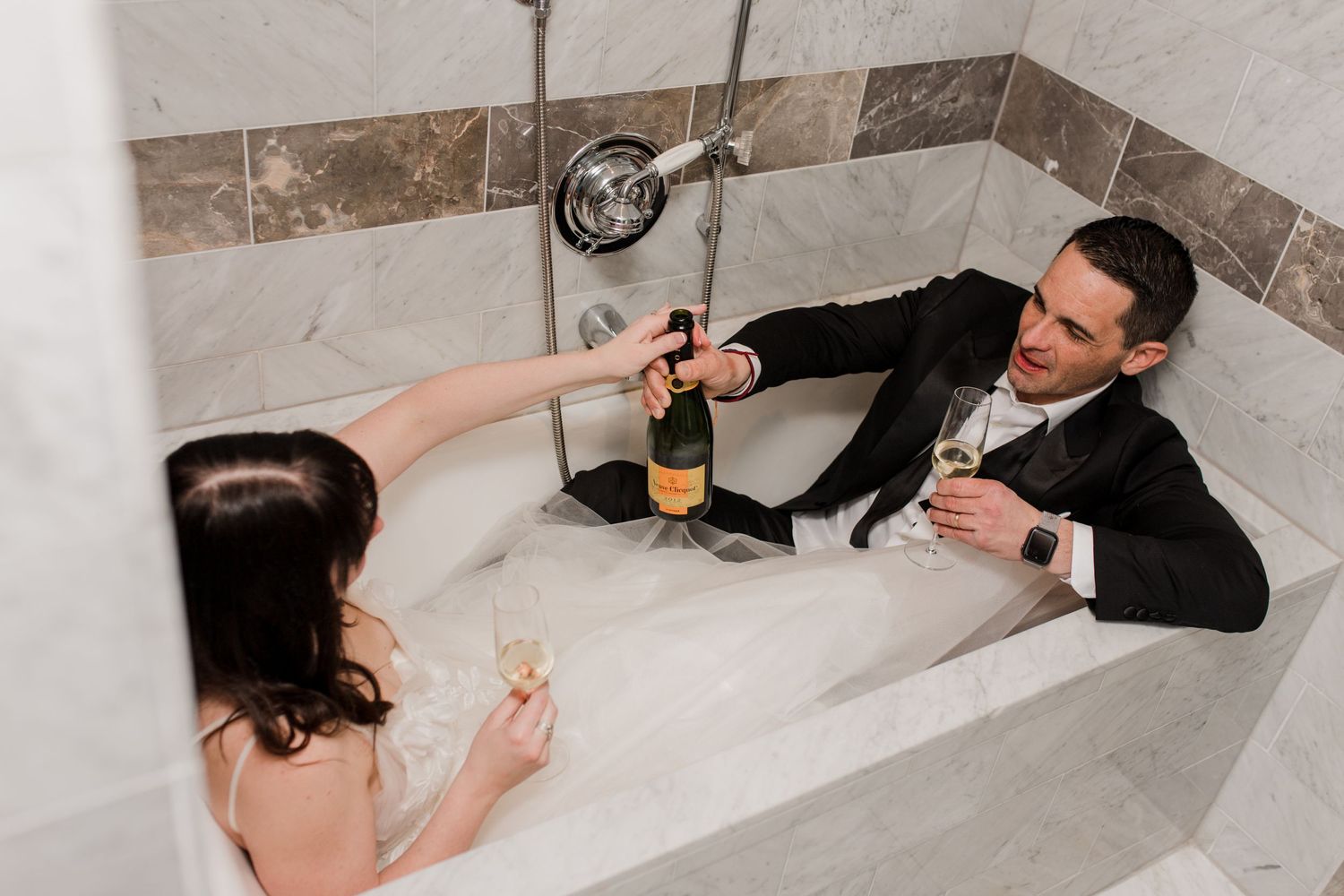 A couple sits facing each other in a bathtub in their wedding attire as they pass a bottle of champagne.