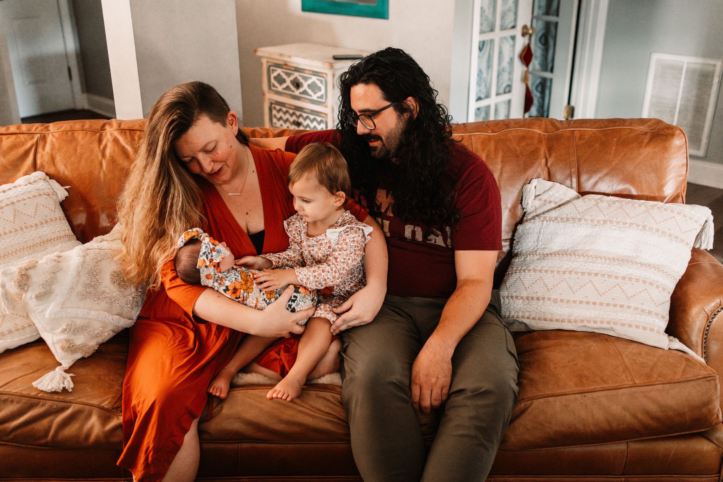 A young family with a toddler sits together on a brown leather couch in a cozy living room.