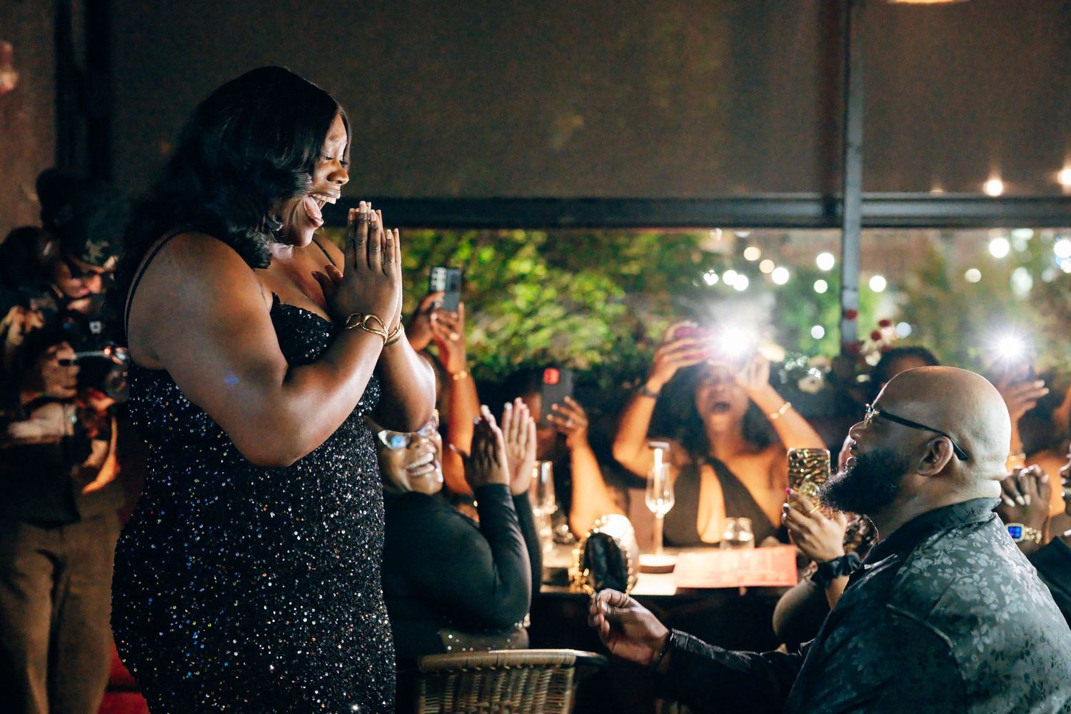 Woman in a sparkling dress covers her mouth in surprise as a kneeling man proposes while diners applaud and film in a candlelit restaurant.