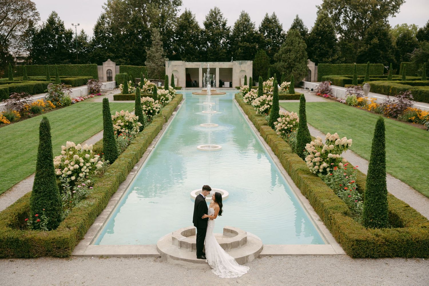 Bride and groom embrace beside a long reflecting pool with fountains in a manicured formal garden with flowers, hedges and a pavilion.