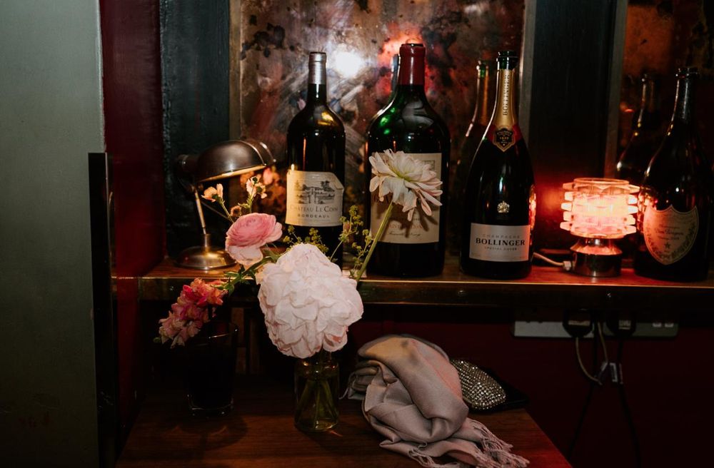 Romantic bar setup with wine bottles, champagne, and pink floral arrangement in warm candlelit ambiance.