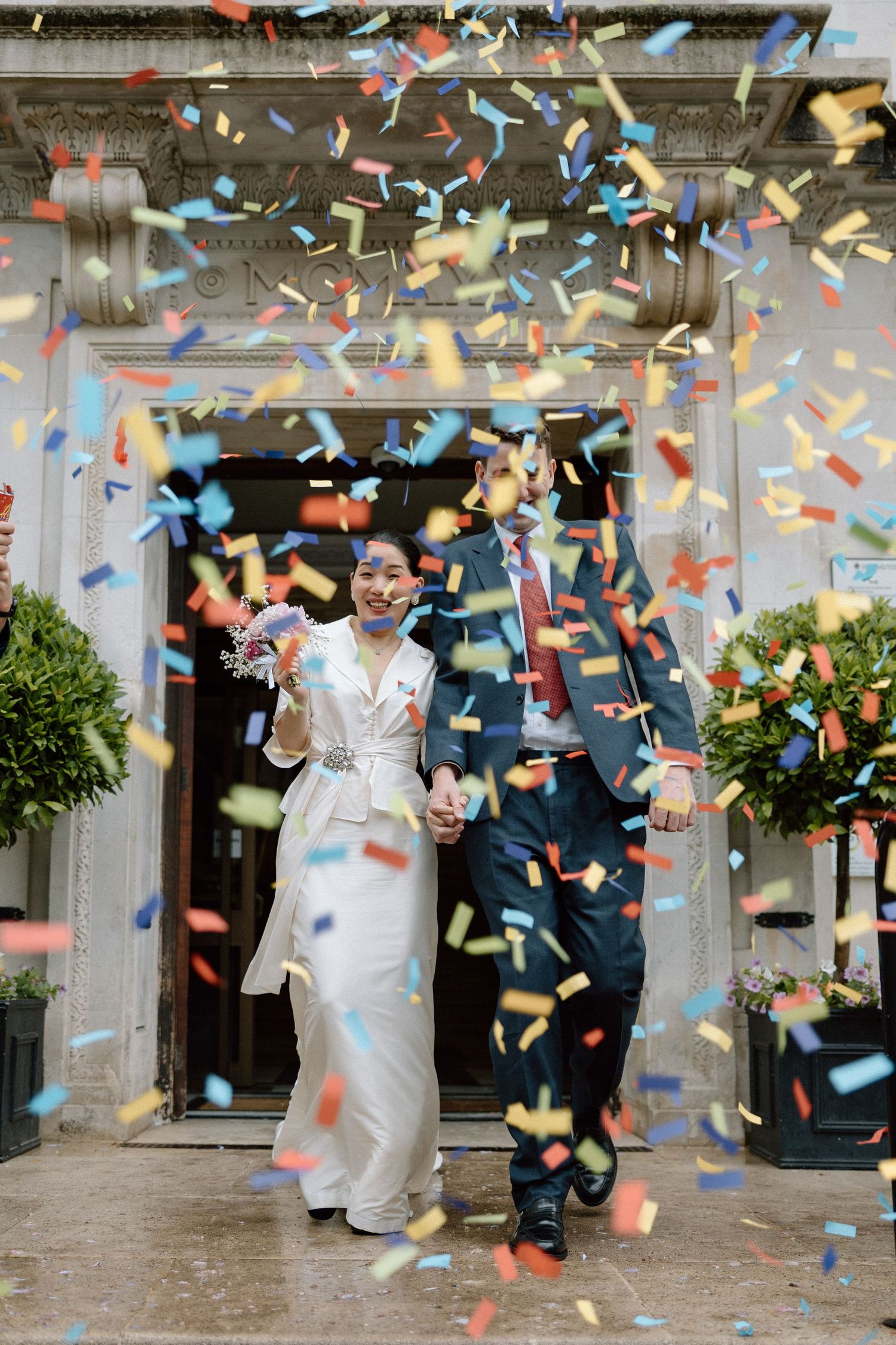 Newlywed couple exits Islington Town Hall holding hands amid colourful confetti shower, bride in white dress, groom in navy.