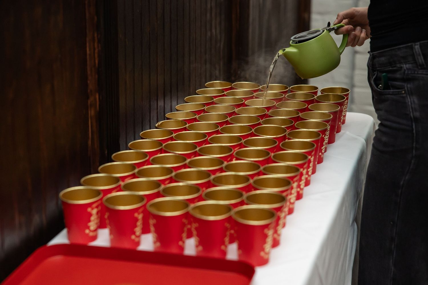 Green teapot pouring hot tea into rows of red and gold cups on a white table.