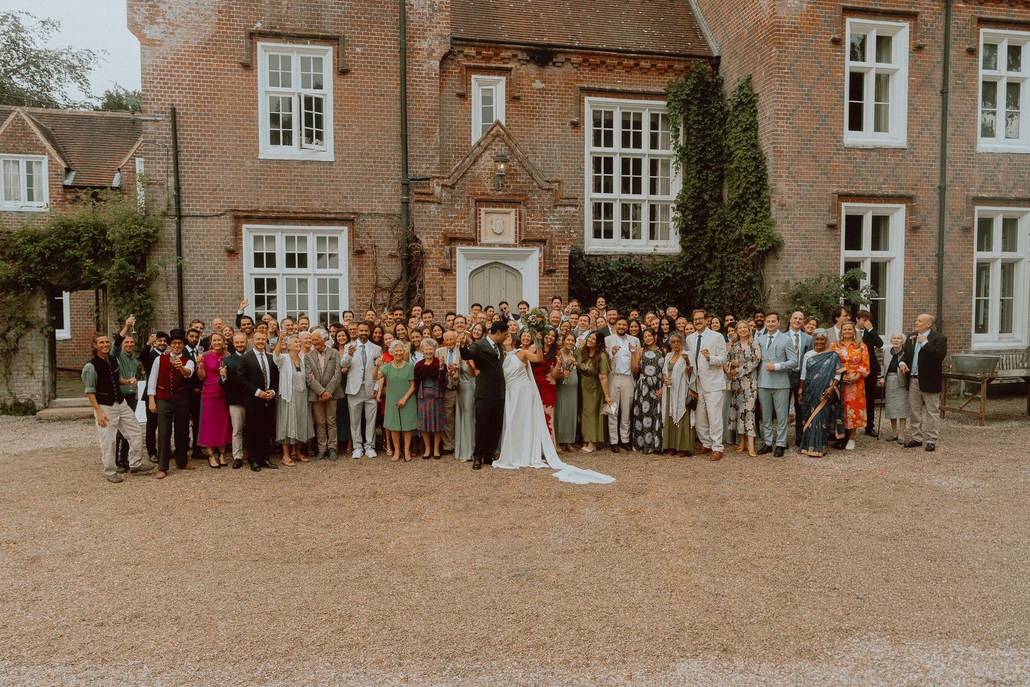 Wedding party group photo with bride and groom surrounded by guests outside a brick manor house.