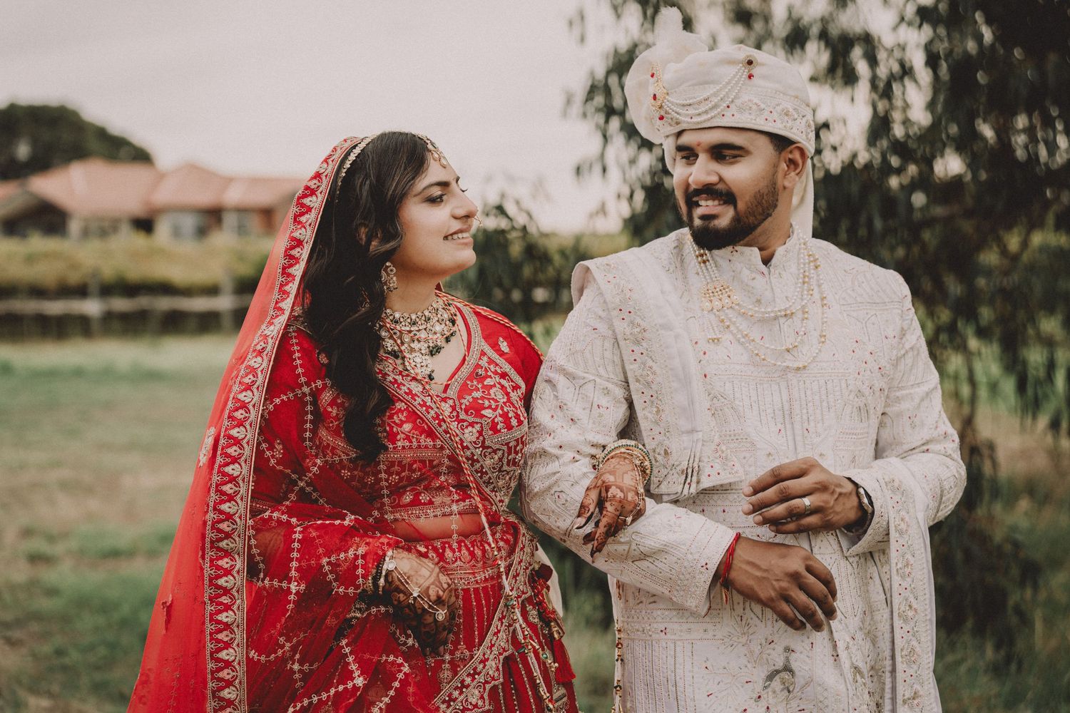 Indian bride in red lehenga and groom in white sherwani smiling at each other during traditional wedding ceremony.