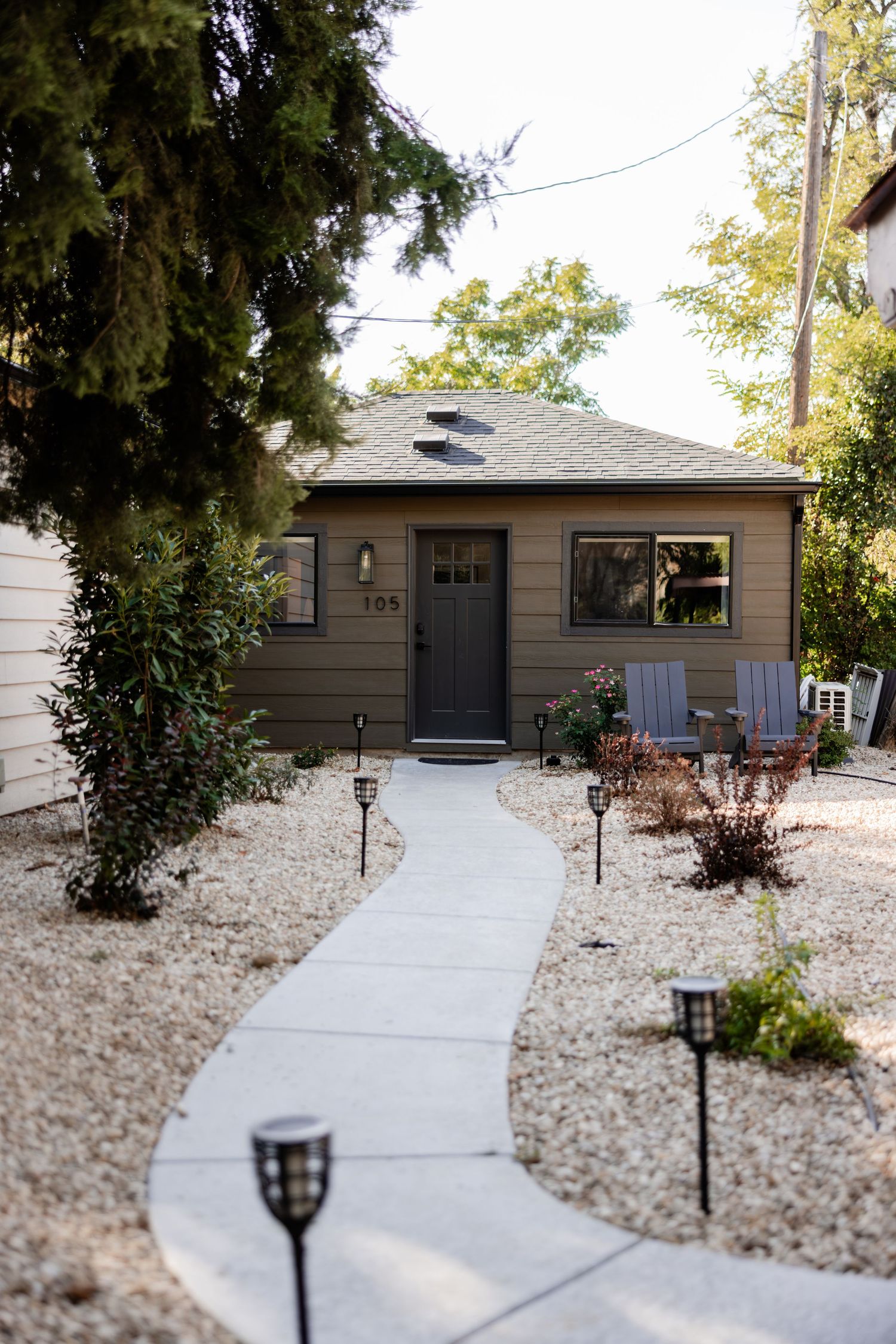 Charming small brown cottage with gravel yard, solar panels, pathway lined with solar lights, and Adirondack chairs.