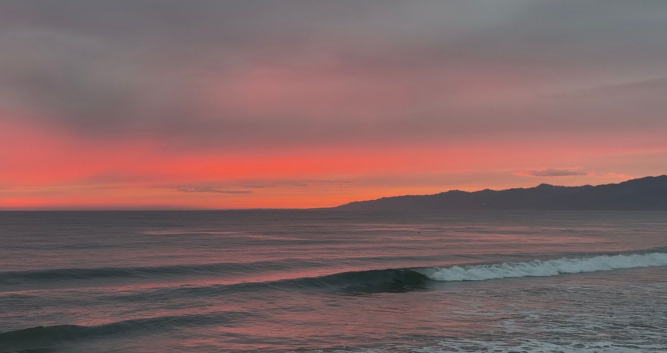 Vibrant pink and red sunset over calm ocean waves with silhouetted mountains in the background.