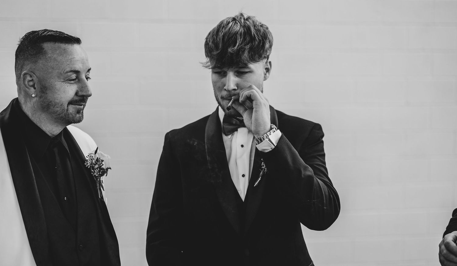 Two men in formal black suits at a wedding, one wearing a boutonniere, the other adjusting his tie in a candid moment.