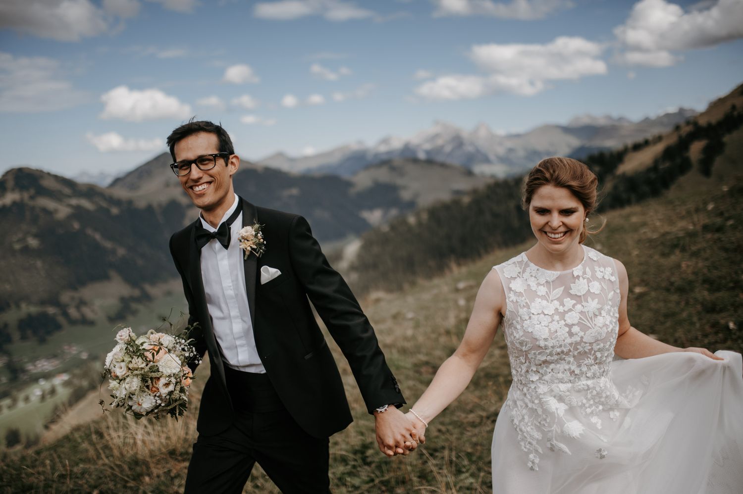 Smiling bride and groom holding hands walking through alpine mountain landscape on wedding day.