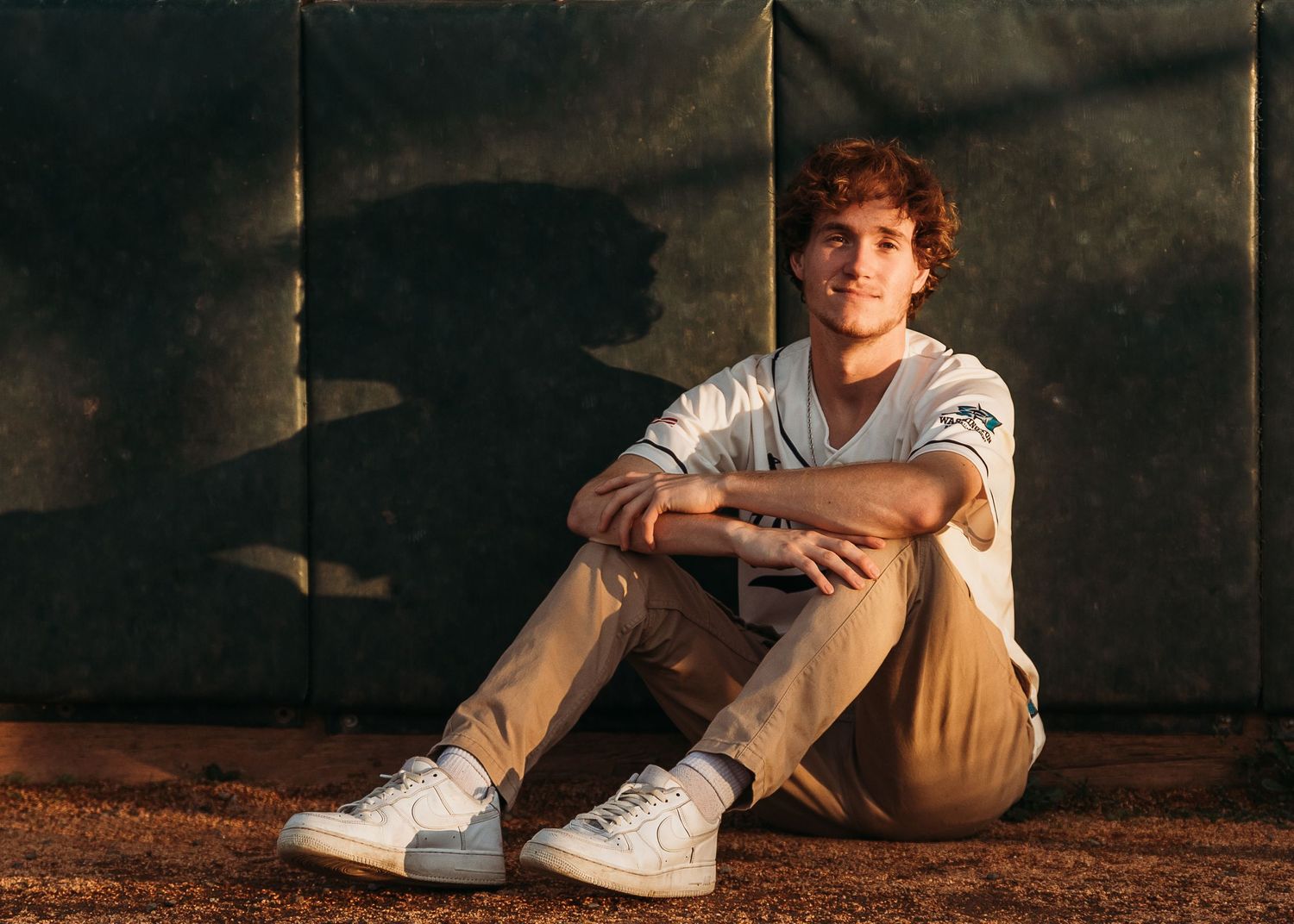 Baseball player poses sitting against wall in stadium wearing white uniform during golden hour.