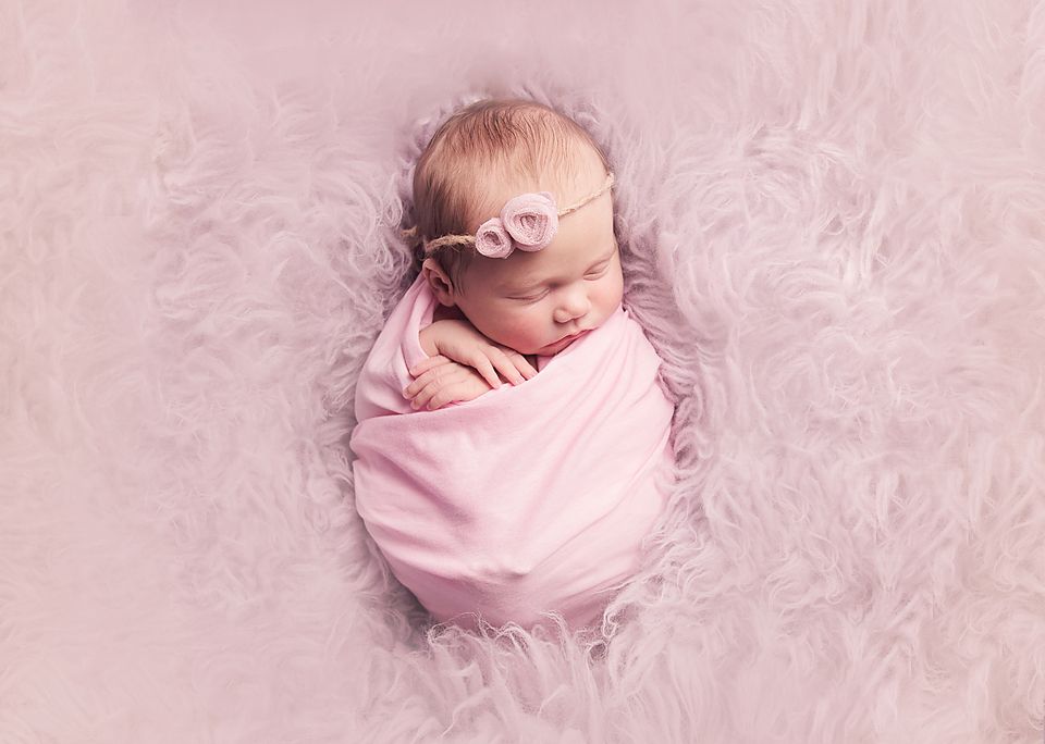 A sleeping newborn baby wrapped in pink swaddle blanket lying on soft purple fur background.