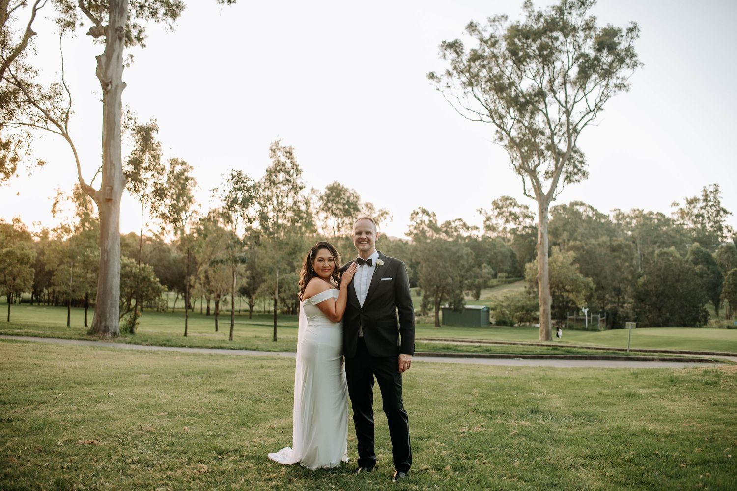 Wedding couple shares an intimate moment on a grassy field with eucalyptus trees at sunset.