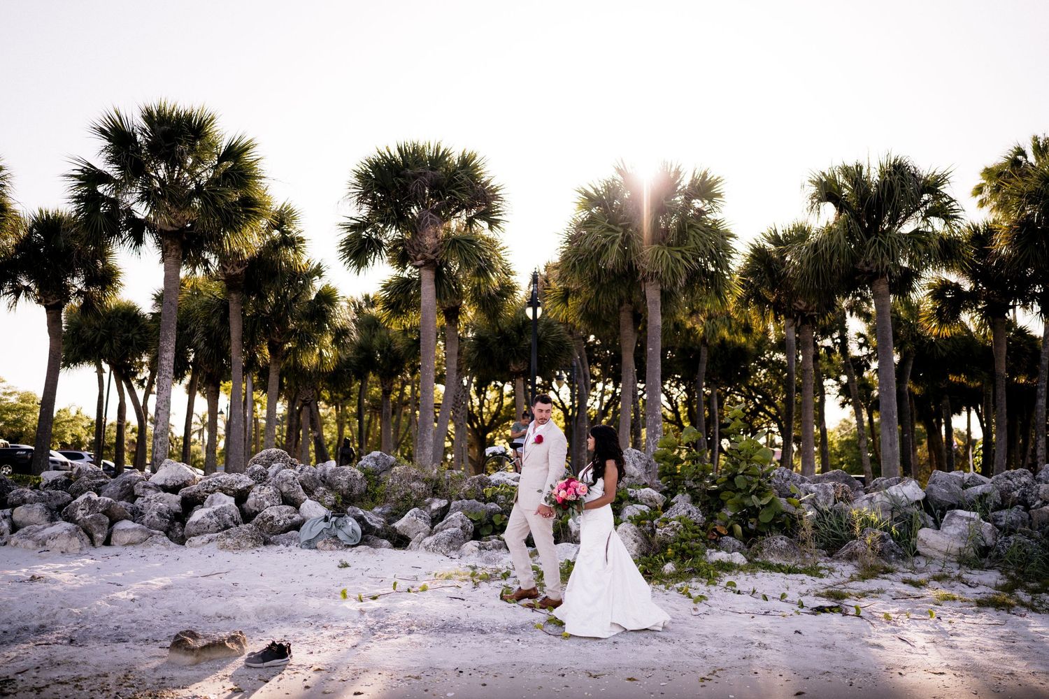 A panoramic beach wedding scene with palm trees and white decor along a sandy shoreline at sunset.