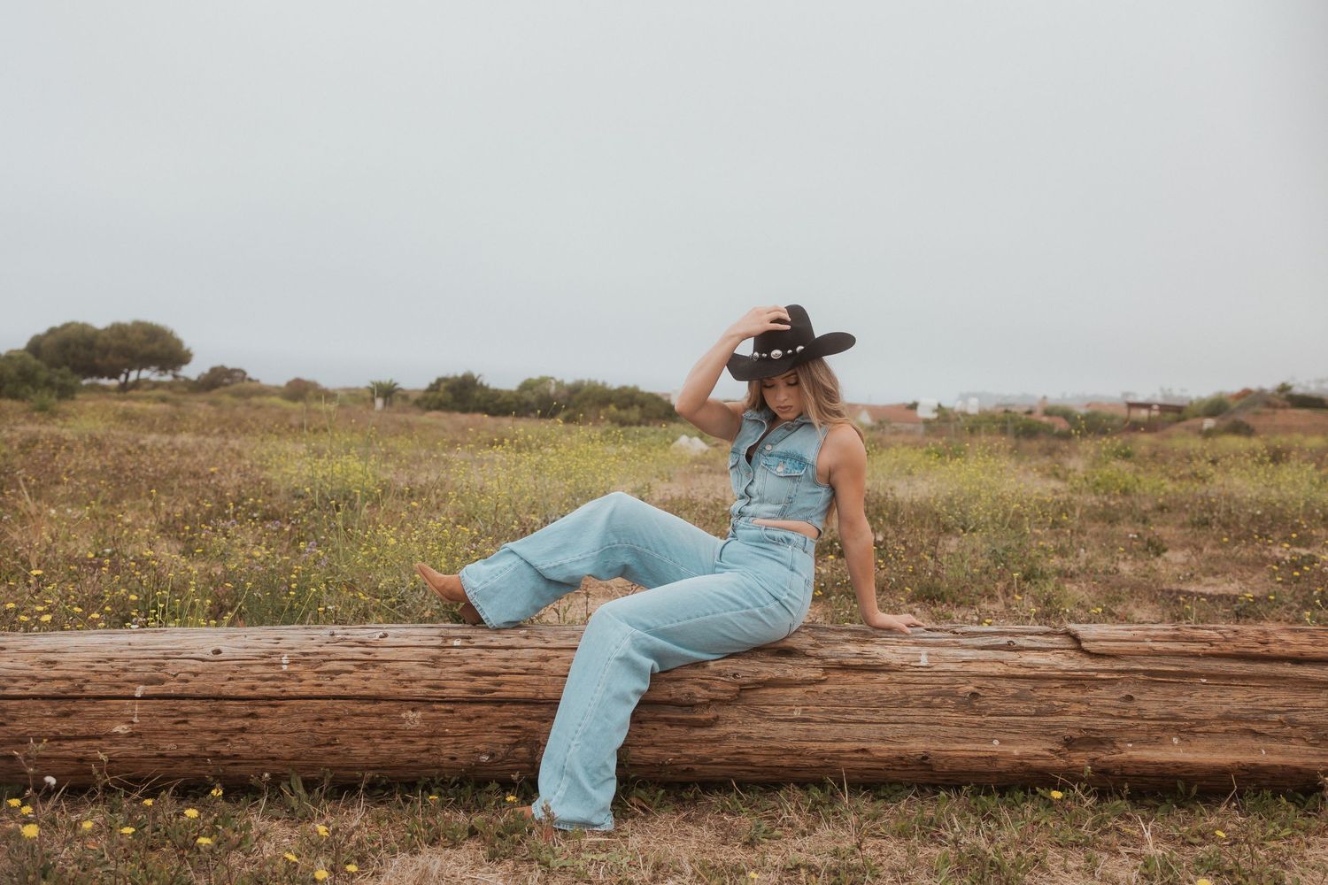 A person in denim overalls sits casually on a rustic log while looking towards the horizon.