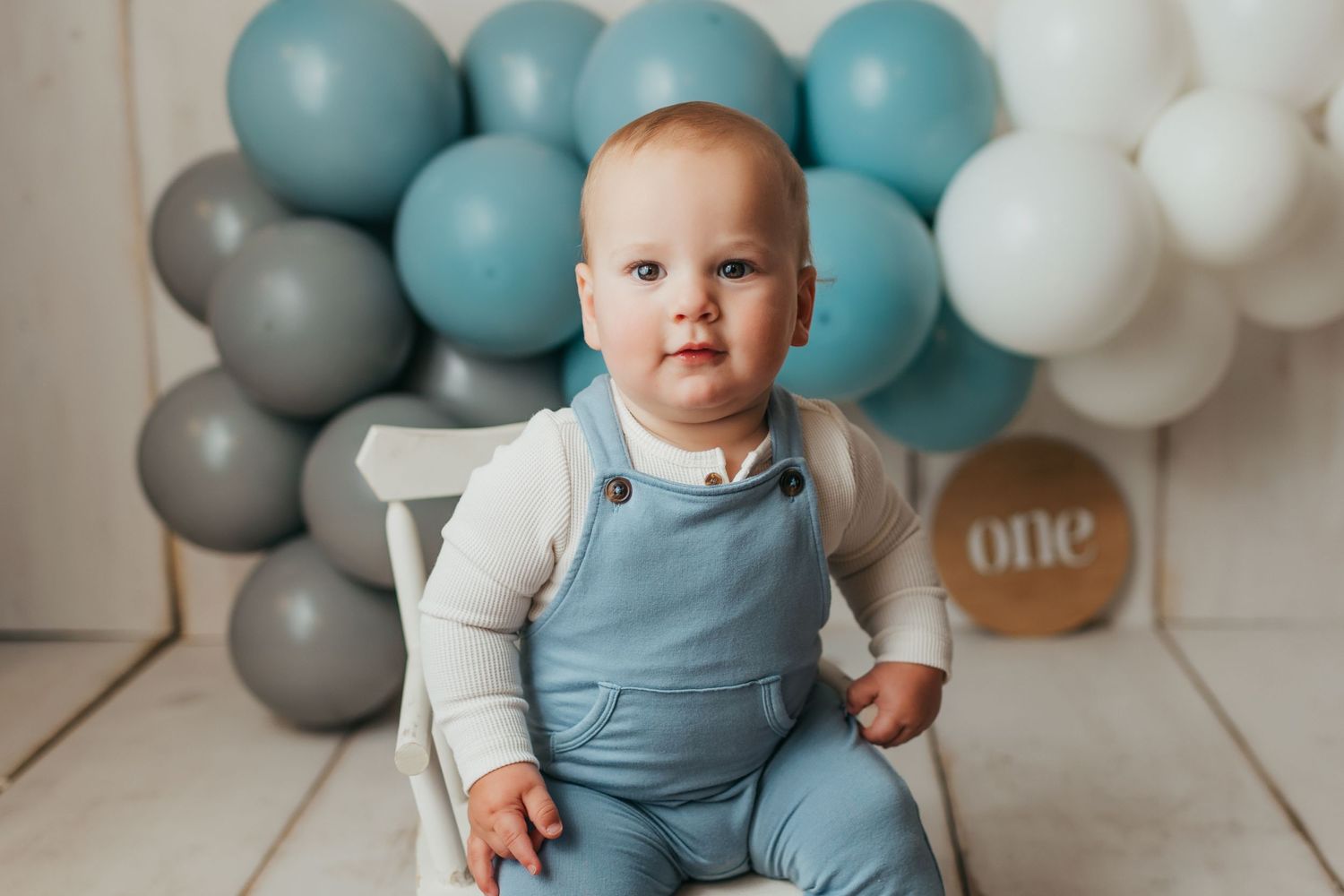 Baby in blue overalls sitting in white chair with balloon arch backdrop.