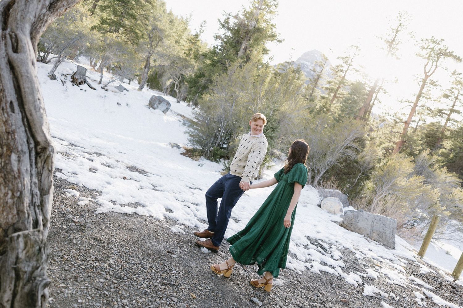 A couple stands together on a snowy mountain trail, with the person in a flowing emerald green dress.
