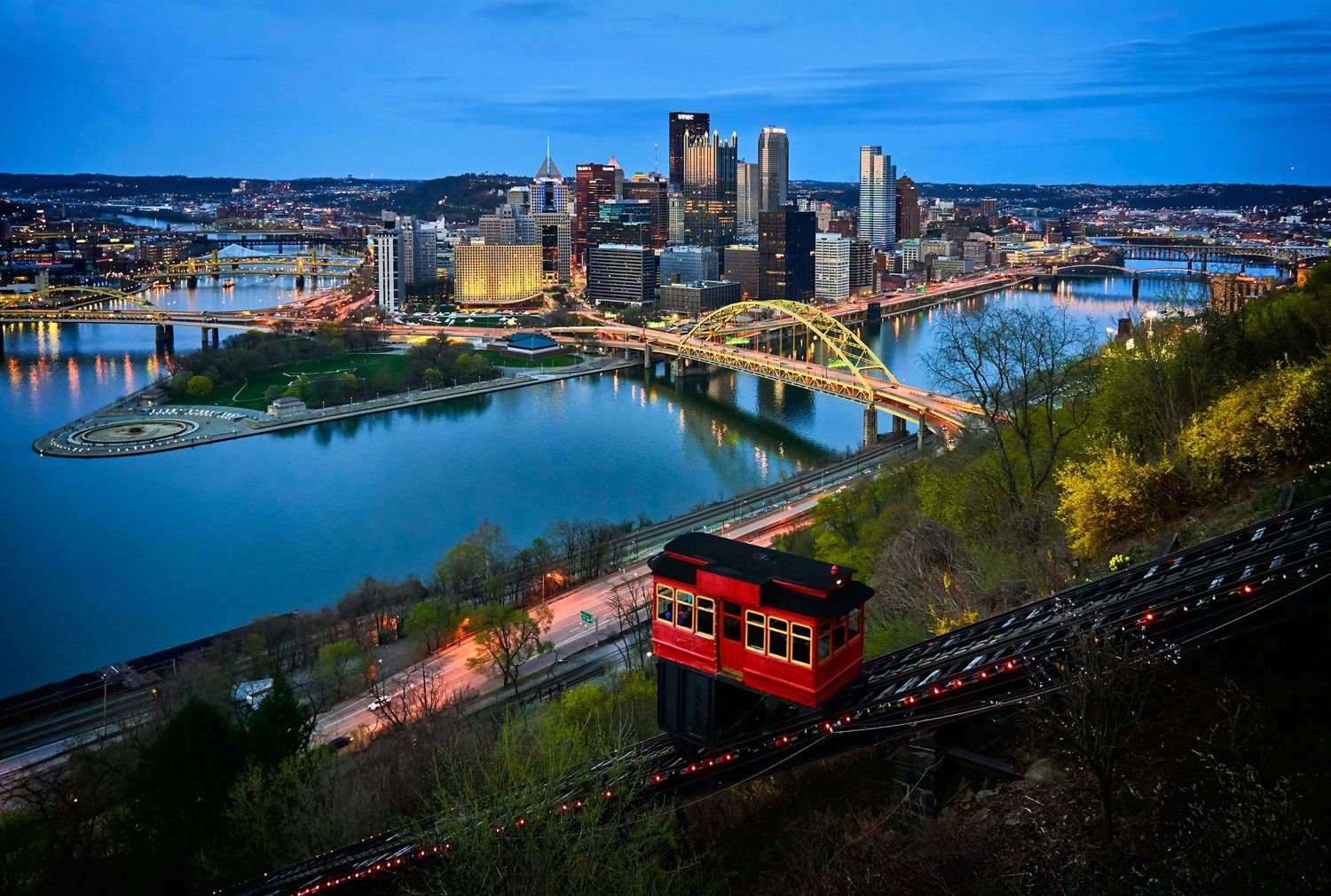 A red Duquesne Incline cable car overlooks downtown Pittsburgh's skyline and three rivers at dusk.