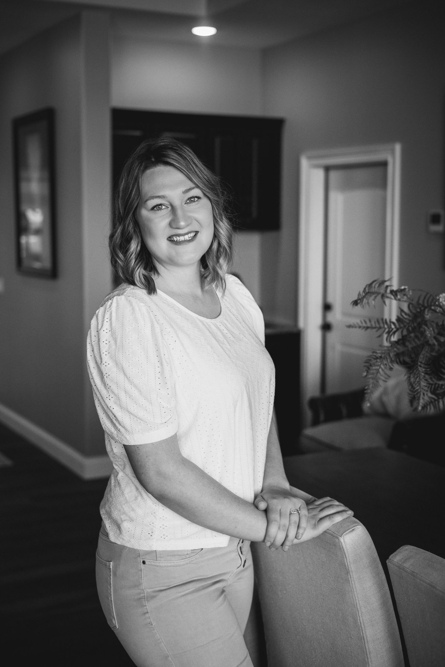 A black and white indoor portrait of someone wearing a white blouse and light-colored pants leaning against furniture.