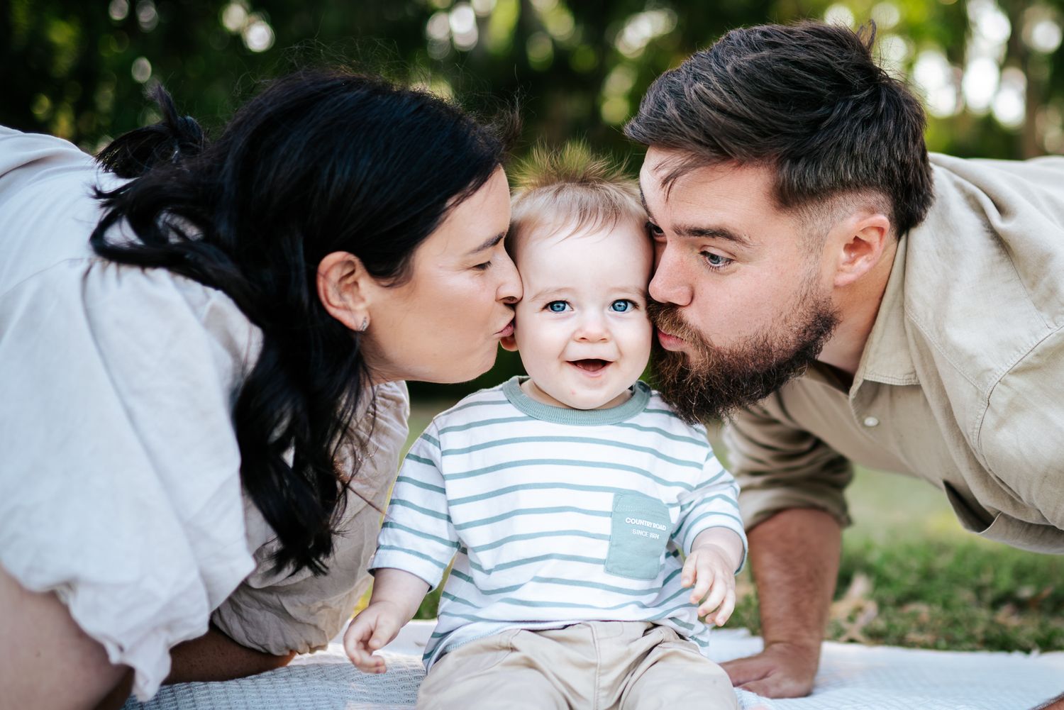 A family sits together on a white blanket in a sunny park with trees in the background taken by Melbourne family photographer.