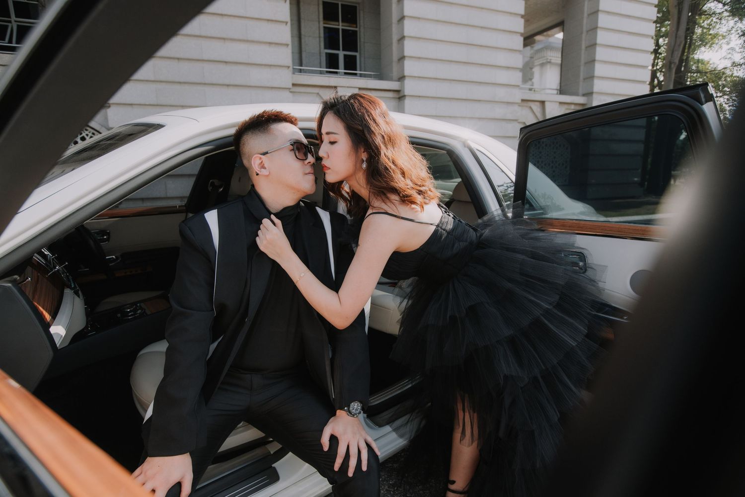 Elegant couple in black formal attire share an intimate moment in luxury car with tan leather interior.