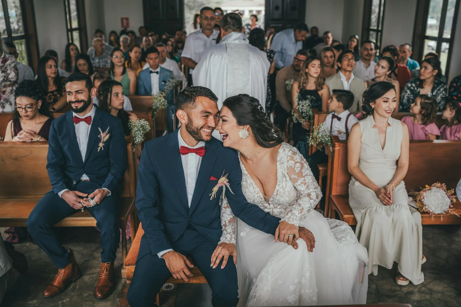 A couple shares an intimate moment while seated together during a wedding ceremony with guests looking on in the background.