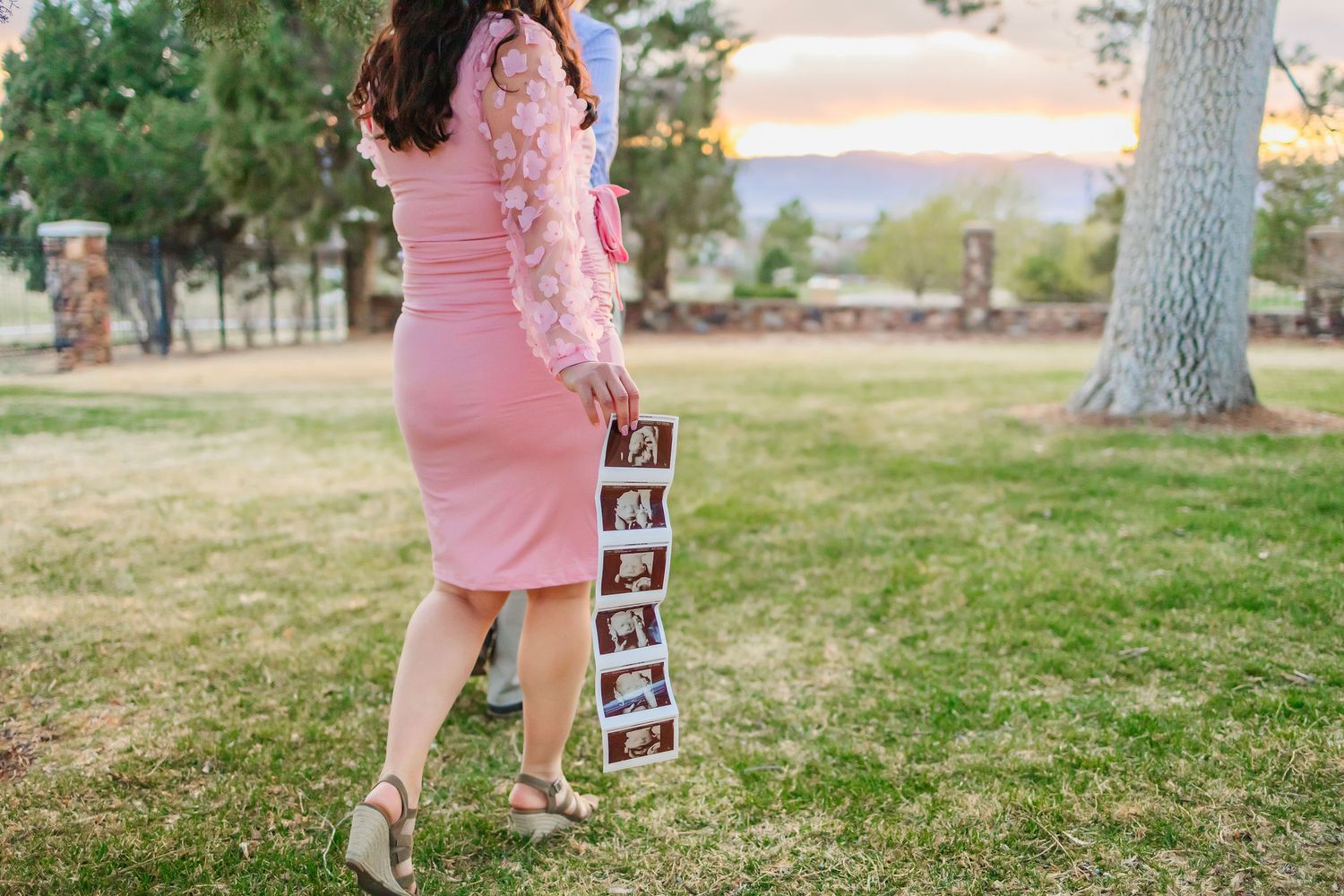 Pregnant woman in pink dress holding ultrasound photos outdoors at sunset in a lush green park.