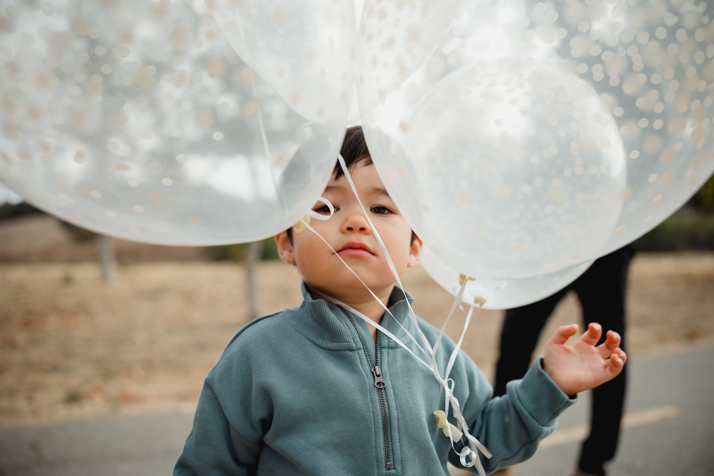 Young child in blue jacket looking up with wonder at large white balloons against a blurred outdoor background.