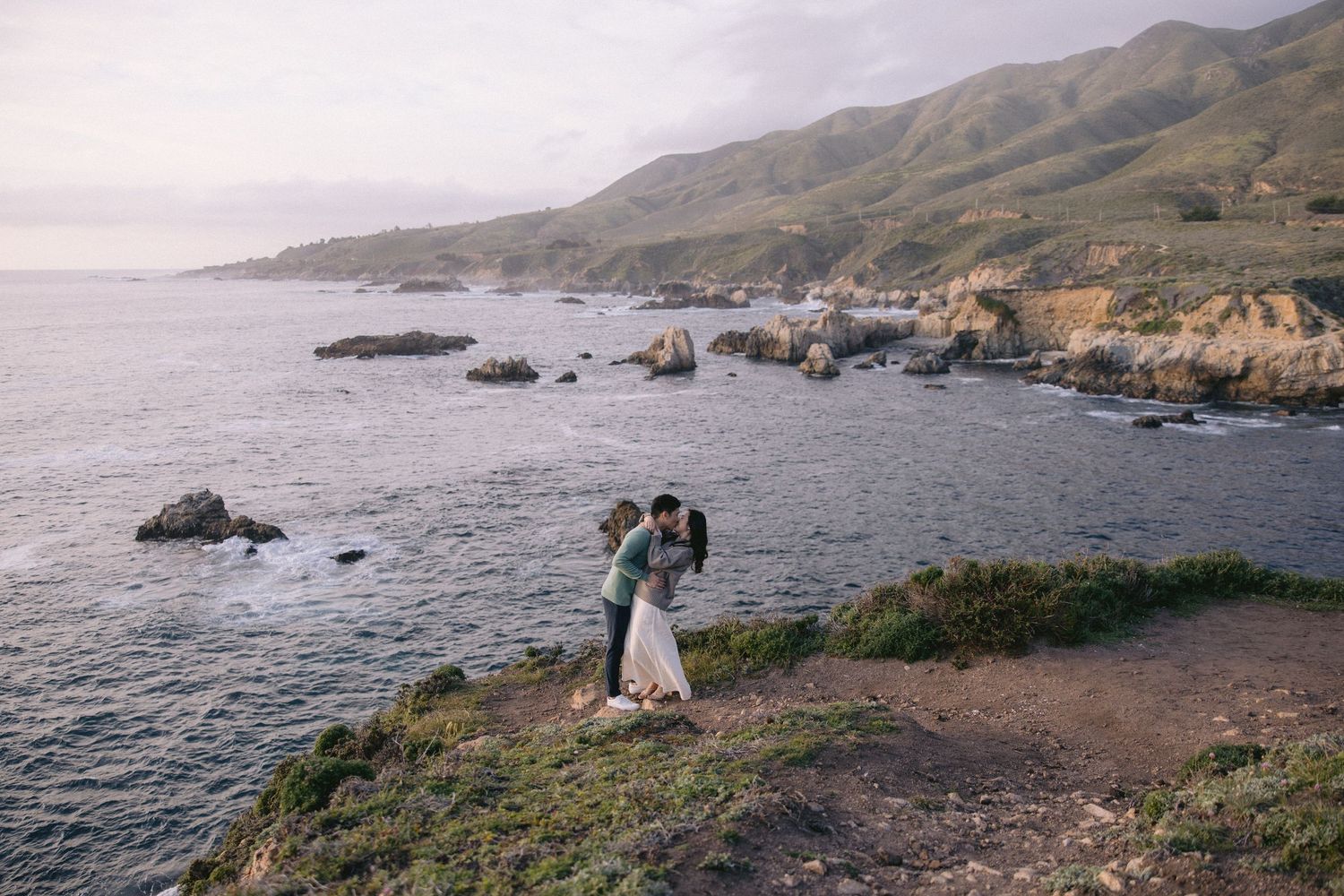 Two figures embrace on a scenic cliff overlooking the rugged Big Sur coastline with rocky outcrops and mountains at sunset.