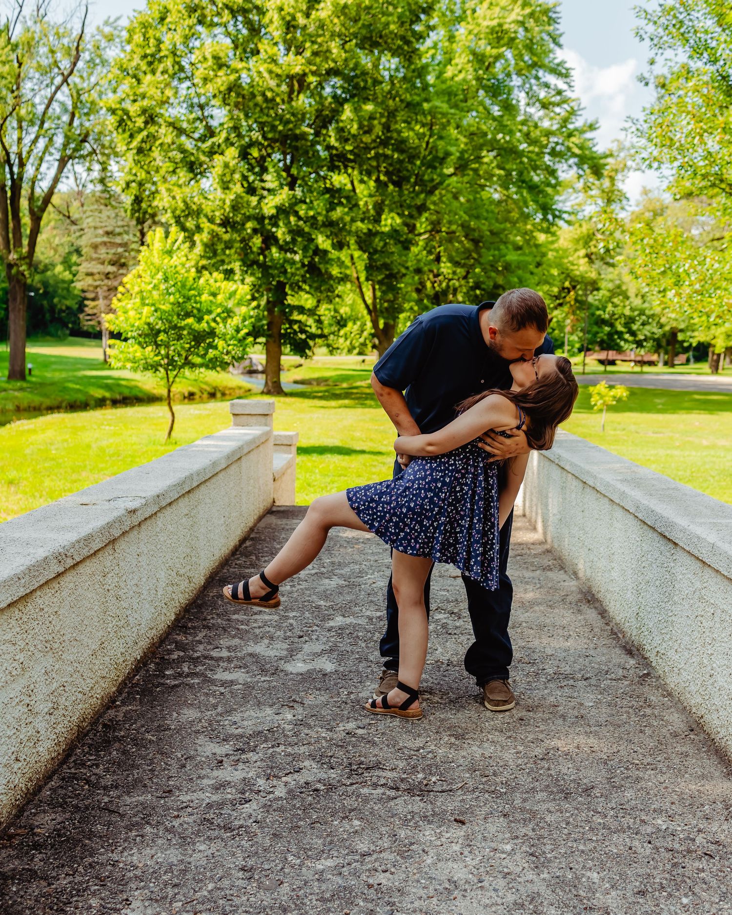 A romantic couple shares a tender moment while embracing on stone steps in a lush green park setting.