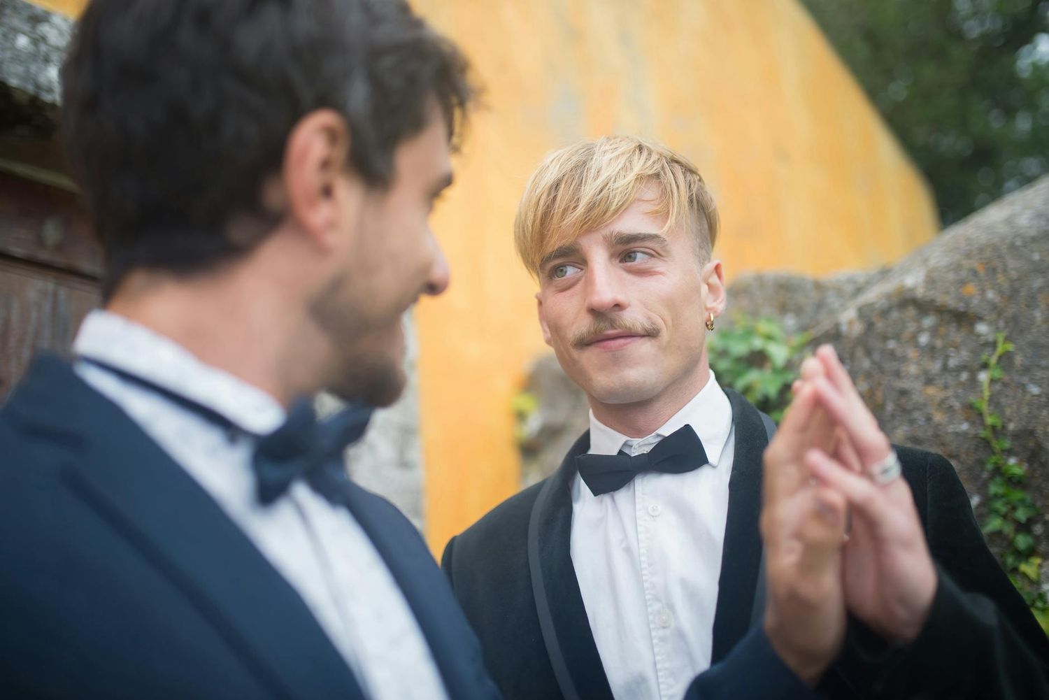 Two people in tuxedos and bow ties share a moment at a formal event against a yellow wall background. LGBT Pittsburgh Wedding Photography