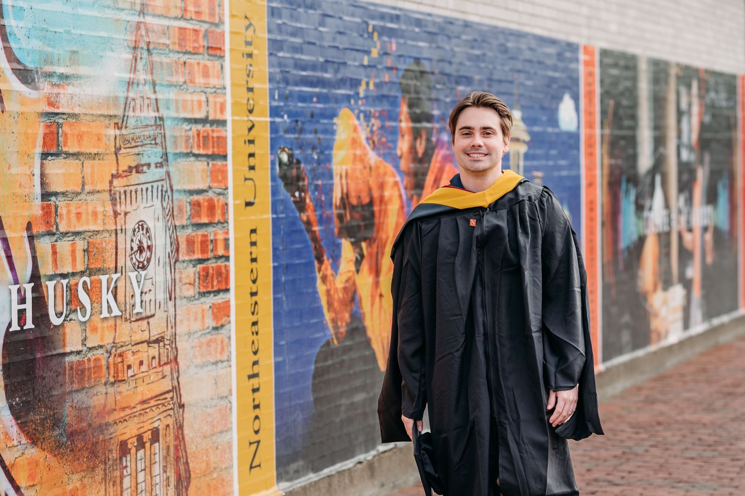 A graduate in academic regalia stands smiling in front of a colorful brick wall mural on an urban street.