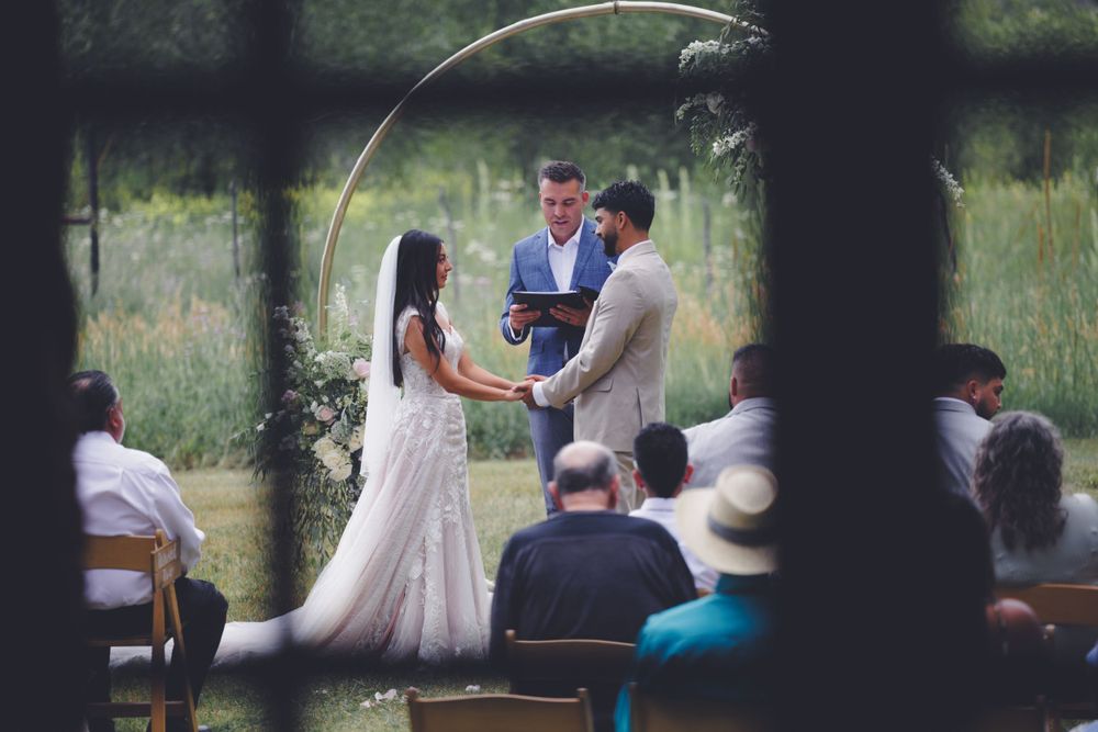 A couple holding hands during an outdoor wedding ceremony, with a lush green background and seated guests.