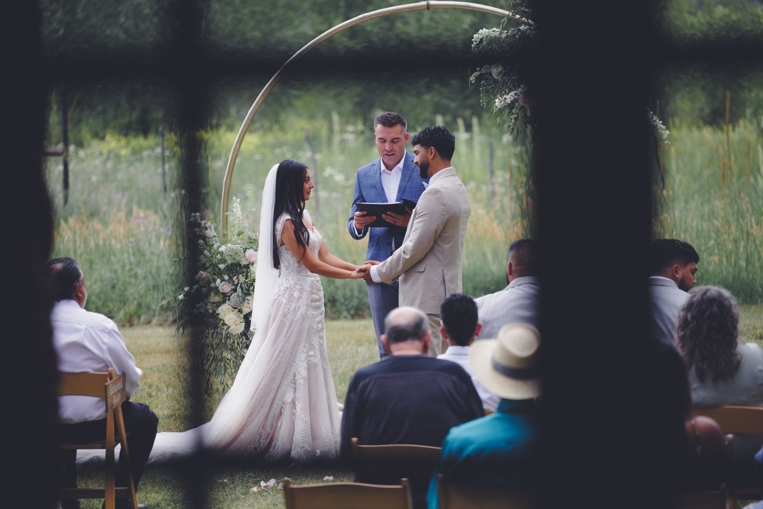 A couple holding hands during an outdoor wedding ceremony, with a lush green background and seated guests.