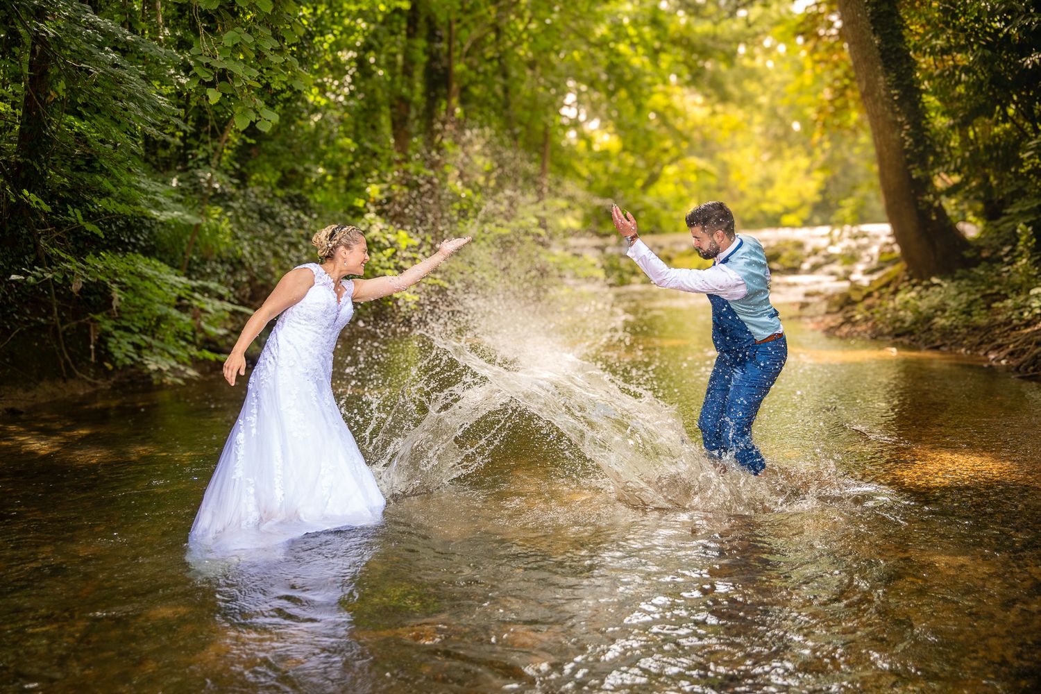 Wedding couple creates splash in forest stream during playful photo shoot.