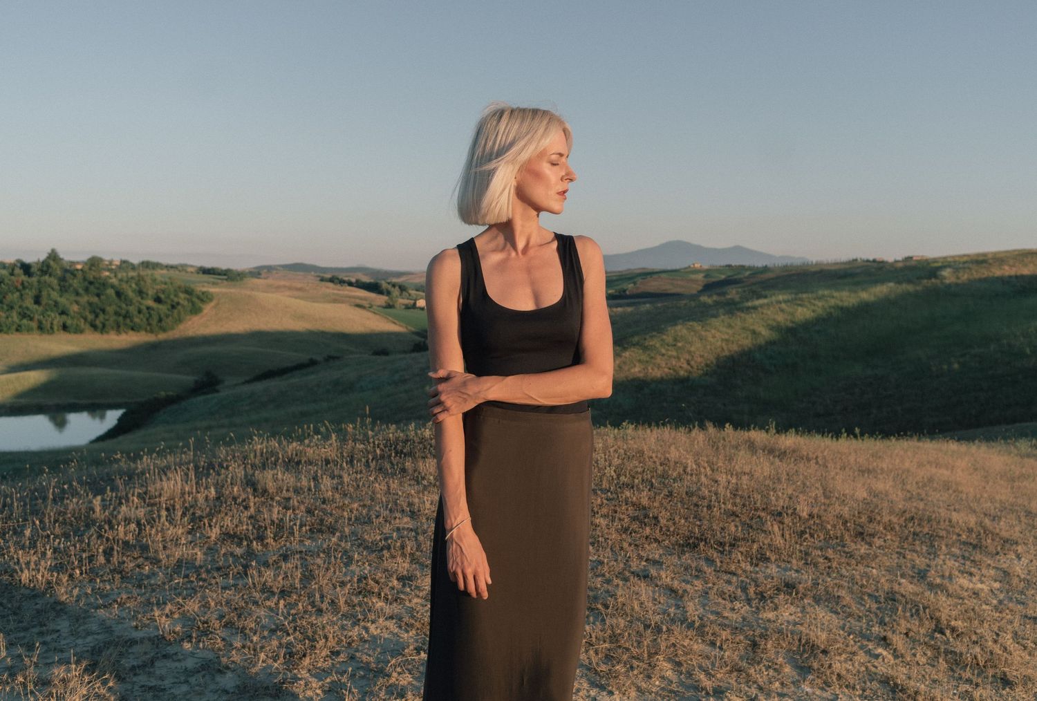 Figure in a long brown dress stands against rolling hills at sunset in Tuscany countryside.