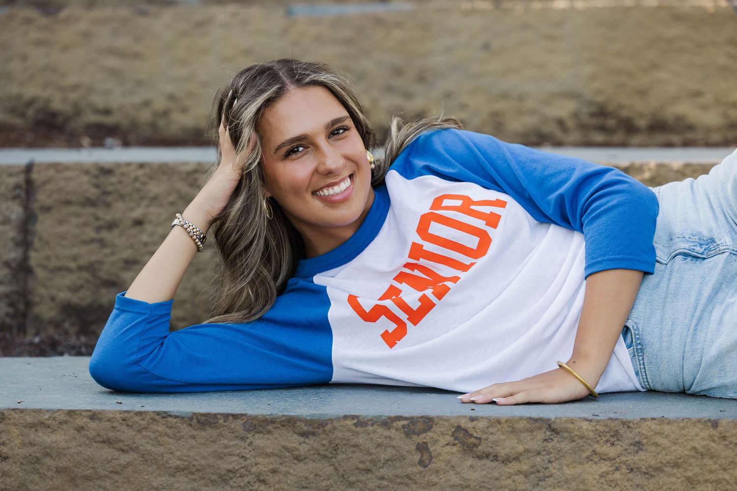 Smiling senior girl in blue and white baseball tee laying on concrete steps for senior portrait photo.