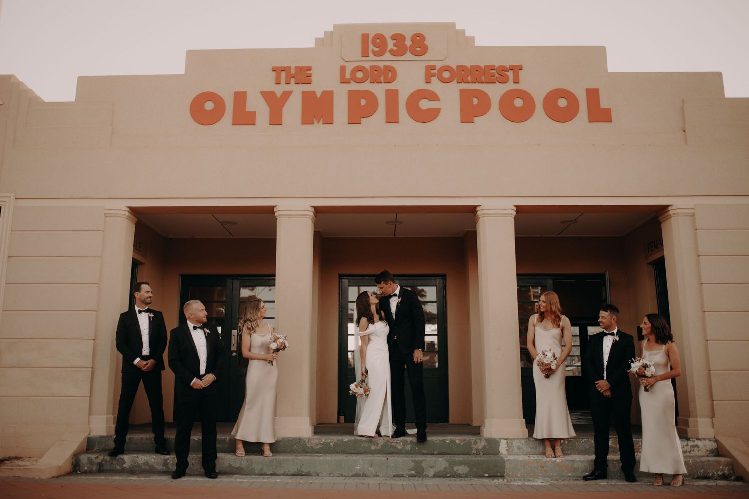 Vintage-style wedding party posed in front of historic 1938 Olympic Pool building.