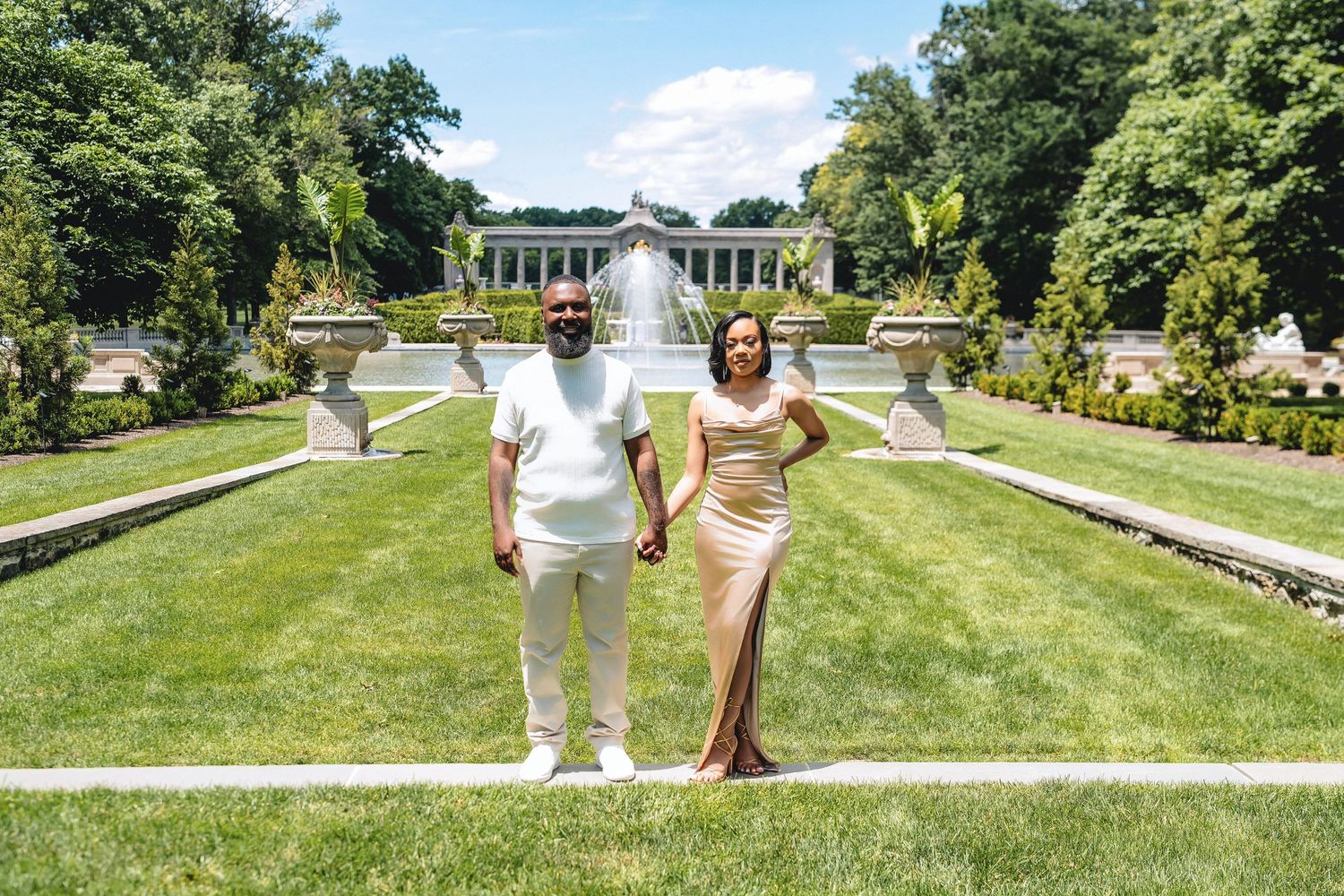 Elegant couple poses in white and gold attire by fountain in formal garden setting.