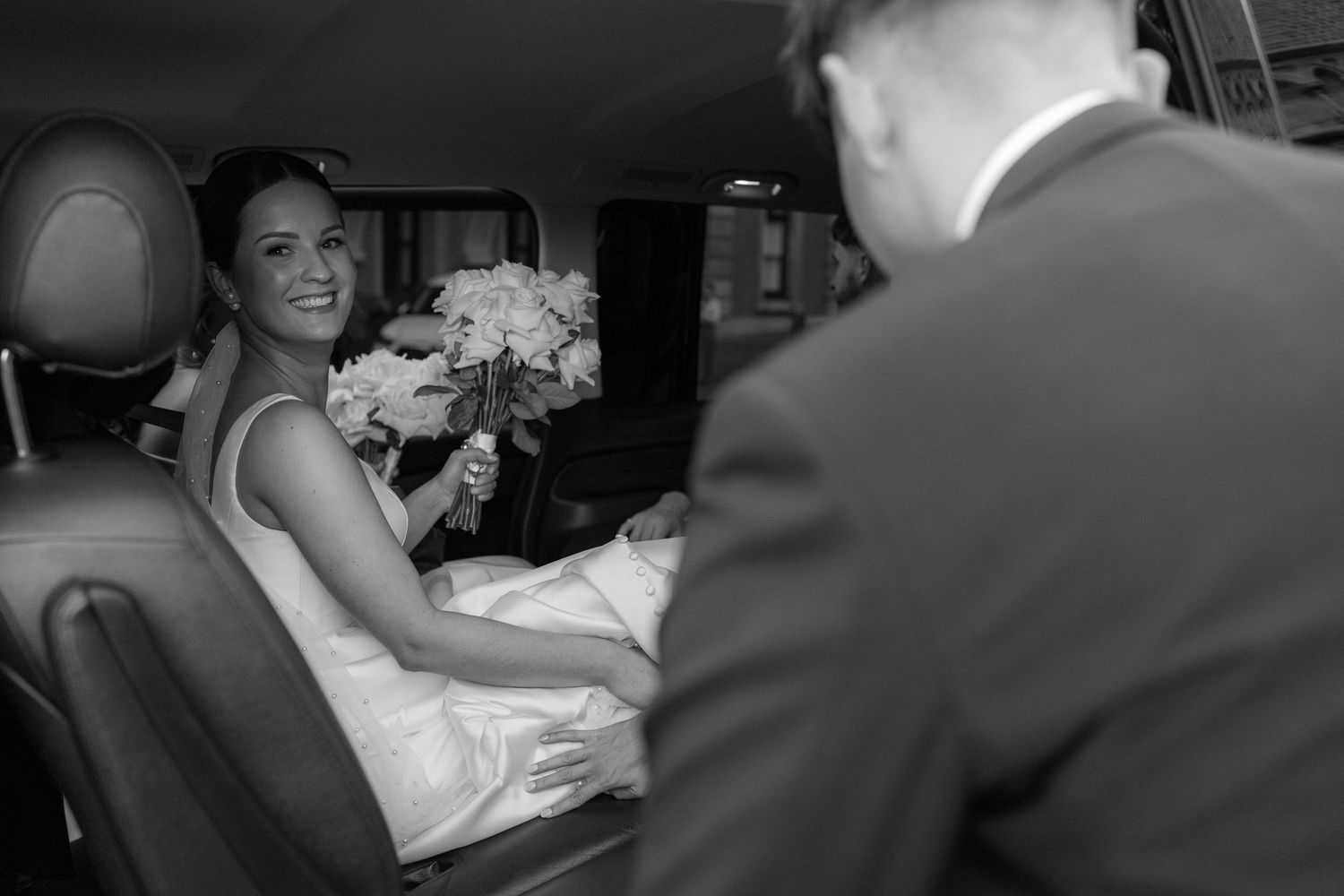 Black and white photo of bride with bouquet sitting in car.
